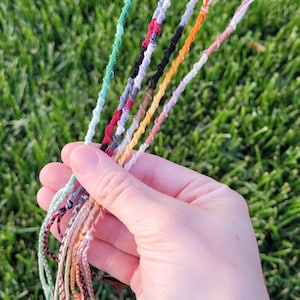 Può includere: Una collezione di braccialetti dell'amicizia colorati realizzati con corda intrecciata. I braccialetti sono di vari colori, tra cui verde, rosso, bianco, nero, arancione e beige.