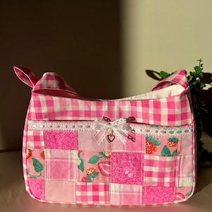 May include: A pink patchwork handbag with a gingham pattern and strawberry print. The bag features a lace trim, a bow, and a heart charm. The bag has a rectangular shape with a top handle.