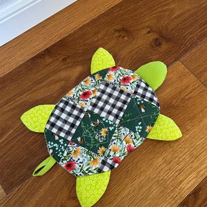 Puede incluir: Un artículo acolchado hecho a mano con forma de tortuga. El caparazón de la tortuga es un patchwork de telas florales, cuadros y verdes lisos. La cabeza y las aletas son de color verde brillante. La tortuga está sobre una superficie de madera.
