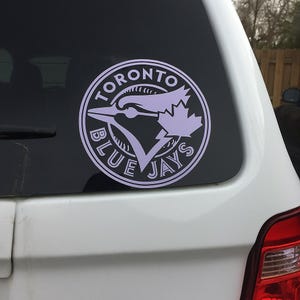 May include: A light purple Toronto Blue Jays logo sticker on a car window. The circular design features a blue jay bird, a maple leaf, and the words "TORONTO" and "BLUE JAYS" in a white font.
