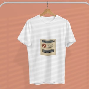 May include: White t-shirt with a graphic of a computer error message that reads "Error" and "art not loaded". The graphic is in shades of gray and beige. The shirt is hanging on a clothing rack.