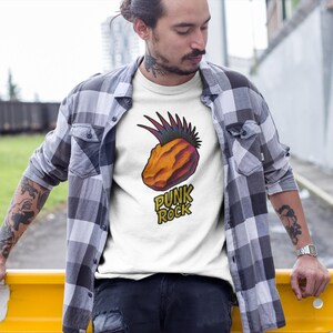Op de afbeelding: Wit T-shirt met een afbeelding van een ananas met een hanenkam en de woorden "PUNK ROCK" in het geel. De persoon draagt een grijs en zwart geruit overhemd.