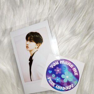 May include: A white photo card with a photo of a person with black hair wearing a white jacket. The photo card is next to a round sticker with a holographic background and the text "You make me feel groovy" in a rainbow font.
