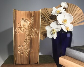 Könnte beinhalten: Ein Buch mit einem 3D-Origami-Fischdesign auf dem Einband. Das Buch steht auf einer braunen Oberfläche mit einer grünen Oberfläche dahinter. Eine blaue Vase mit weißen Blumen steht rechts im Bild. Die Vase steht auf einem Stapel Bücher.