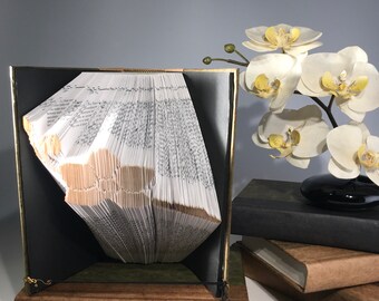 Könnte beinhalten: Eine Buchkunstskulptur in Form eines Vogels, hergestellt aus den Seiten eines Buches. Die Skulptur wird auf einem braunen Buch mit einer daran befestigten schwarzen Schnur präsentiert. Die Skulptur ist von weißen Orchideen und grauen Steinen umgeben.