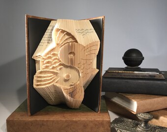 Könnte beinhalten: Eine Buchkunstskulptur eines Yin-Yang-Symbols, hergestellt durch Falten der Seiten eines Buches. Die Skulptur wird auf einem braunen Sockel ausgestellt und ist von anderen Büchern und einem antiken Tintenfass umgeben.