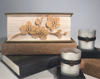 Könnte beinhalten: Ein Buch mit braunem Einband und grünem Rücken, mit einem Papier-Schnitt-Design einer Orchideenblüte auf der Vorderseite. Das Buch liegt auf zwei weiteren Büchern, einem braunen und einem schwarzen. Zwei Kerzen mit schwarzen Bändern und Metallakzenten stehen vor den Büchern.