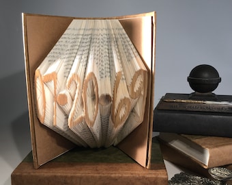 Könnte beinhalten: Eine Buchkunstskulptur der Zahl "07-20-69", gefaltet aus den Seiten eines Buches. Die Skulptur wird auf einem Holzsockel präsentiert und ist von anderen Büchern und Schreibutensilien umgeben.