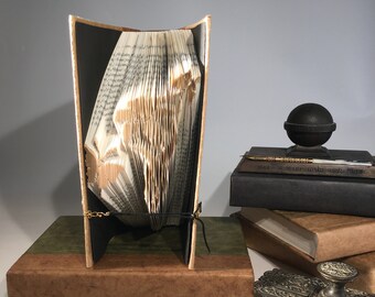 Könnte beinhalten: Eine Buchkunstskulptur, die zu einer dreidimensionalen Form gefaltet ist, wird auf einem braunen und grünen Buch ausgestellt. Die Skulptur besteht aus einem Buch mit schwarzem Einband und ist mit einem schwarzen Band gebunden. Die Skulptur wird auf einem Tisch mit einem schwarzen Tintenfass und einem Stapel Bücher ausgestellt.