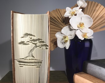 Könnte beinhalten: Eine Buchkunstskulptur eines Bonsai-Baums, gefaltet aus den Seiten eines Buches. Die Skulptur wird auf einem braunen Sockel präsentiert und steht neben einer blauen Vase mit weißen Orchideen und einem braunen Fächer.