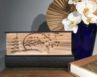 Könnte beinhalten: Ein Buch mit einem geschnitzten Design eines Baumes und eines Mondes auf dem Einband. Das Buch liegt auf anderen Büchern, und im Hintergrund steht eine Vase mit weißen Orchideen.