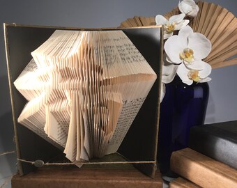 Könnte beinhalten: Eine aus einem Buch gefaltete 3D-Origami-Skulptur eines Baumes, die auf einem Holzsockel präsentiert wird. Die Skulptur ist in einem schwarzen Rahmen gerahmt und steht auf einem Tisch mit einem Stapel Bücher und einer Vase mit weißen Orchideen.