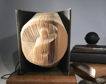 Könnte beinhalten: Eine Buchskulptur eines Yin-Yang-Symbols, gefaltet aus den Seiten eines Buches. Die Skulptur wird auf einem Holzsockel präsentiert und ist von anderen Büchern und einem dekorativen Tintenfass umgeben.
