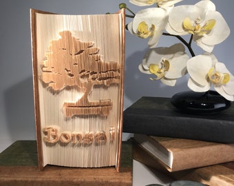 Könnte beinhalten: Ein einzigartiges Buchkunstwerk mit einem geschnitzten Bonsai-Baum-Design. Das Wort "Bonsai" ist ebenfalls sichtbar. Die Kanten sind kupferfarben. Das Kunstwerk wird mit anderen Büchern und einer weißen Orchidee präsentiert, was eine ruhige Atmosphäre schafft.