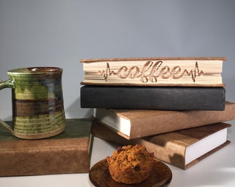 Könnte beinhalten: Ein Stapel Bücher mit einem braunen ledergebundenen Buch obenauf. Das Buch hat das Wort "coffee" mit einer Herzschlaglinie in die Abdeckung geschnitzt. Eine grüne und braune Keramiktasse und ein Muffin stehen auf einem Tisch vor den Büchern.