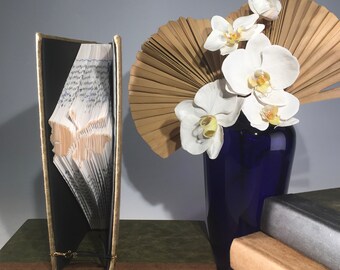 Könnte beinhalten: Eine Buchskulptur in Form eines Vogels, mit gefalteten Seiten, um die Form zu erzeugen. Die Skulptur steht auf einem braunen Buch, neben einem grünen Buch. Eine blaue Vase mit weißen Orchideen und einem getrockneten Fächer aus Palmblättern steht im Hintergrund.