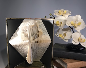 Könnte beinhalten: Eine Buchkunstskulptur eines Baumes, gefaltet aus den Seiten eines Buches, auf einem Holzsockel ausgestellt. Die Skulptur ist geöffnet und die Seiten sind aufgefächert, um die Form eines Baumes zu bilden. Die Skulptur ist von weißen Orchideen und einem Stapel Büchern umgeben.
