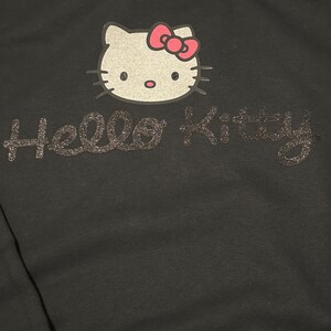Op de afbeelding: Zwarte hoodie met een Hello Kitty-ontwerp in zilveren glitter. De Hello Kitty-afbeelding heeft een roze strik.