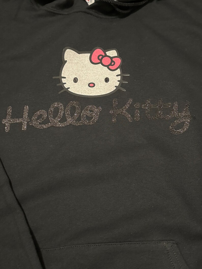 Op de afbeelding: Zwarte hoodie met een Hello Kitty-ontwerp in zilveren glitter. De Hello Kitty-afbeelding heeft een roze strik.