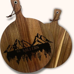 May include: Two round wooden cutting boards with handles. One board features a mountain and forest design with the text "THE MILLERS" in black. The boards have a natural wood grain finish and a leather loop for hanging.
