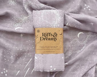 Puede incluir: Una manta de muselina de color morado claro con un estampado celestial blanco de estrellas, lunas y soles. Una banda de papel marrón envuelve el centro con el texto Riffs & Dreams, 70% bambú, 30% algodón orgánico y 120cm x 120cm.