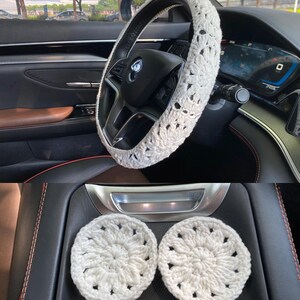 May include: White crocheted steering wheel cover and two matching coasters. The steering wheel cover has a floral design. The coasters are round and have a similar floral design.