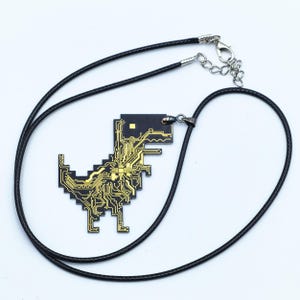 May include: A black necklace with a gold circuit board dinosaur pendant. The necklace is made of a black cord and has a silver clasp.