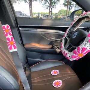 May include: A car interior with a white and pink crocheted steering wheel cover, a white and pink crocheted seatbelt cover, and two white and pink crocheted flower-shaped seat cushions.