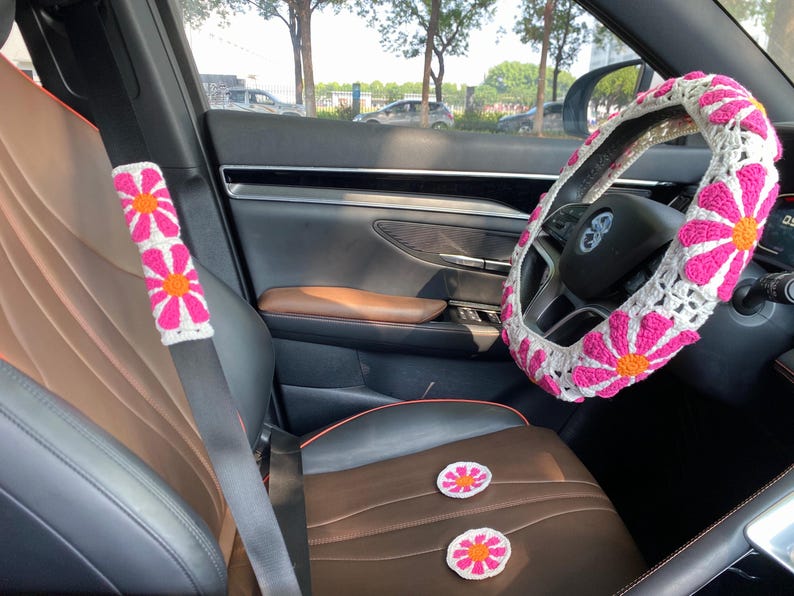 May include: A car interior with a white and pink crocheted steering wheel cover, a white and pink crocheted seatbelt cover, and two white and pink crocheted flower-shaped seat cushions.