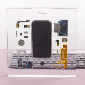 May include: A clear acrylic display case with a disassembled iPhone inside. The phone's screen, circuit boards, and other components are visible. The case is sitting on a gray keyboard.