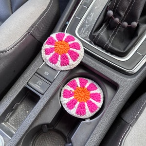 May include: Two crocheted flower-shaped cup coasters in pink and orange. The coasters are sitting in the cup holders of a car.