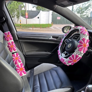 May include: A car interior with pink and white crocheted seatbelt covers and a steering wheel cover with a daisy pattern.