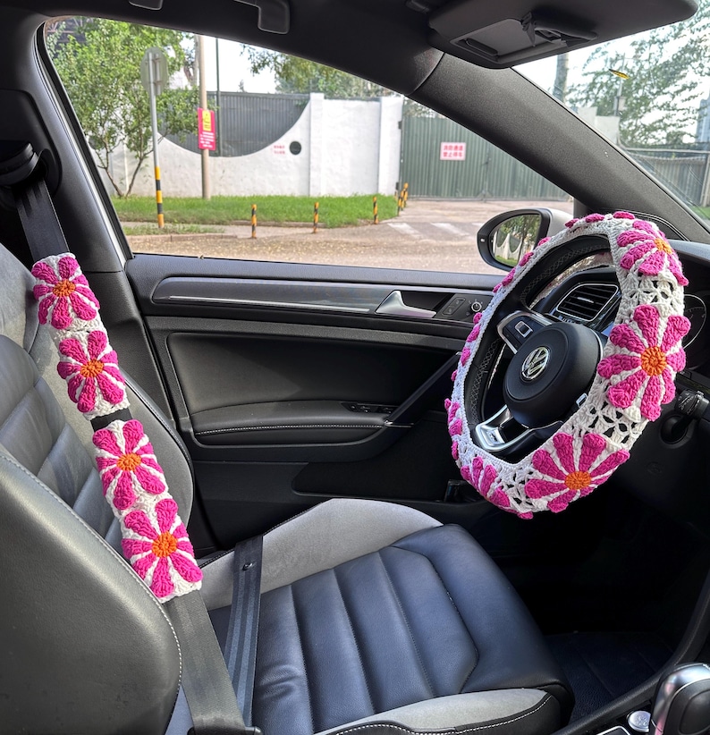 May include: A car interior with pink and white crocheted seatbelt covers and a steering wheel cover with a daisy pattern.