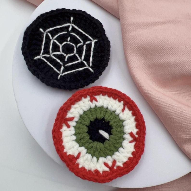 Op de afbeelding: Twee gehaakte Halloween-onderzetters. Een onderzetter is zwart met een wit spinnenwebontwerp. De andere onderzetter is rood met een wit en groen oogontwerp.