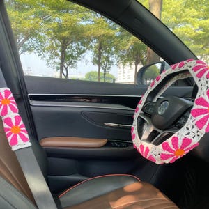 May include: A white crocheted steering wheel cover with pink flowers. The cover is installed on a car's steering wheel. The car's interior is brown leather.