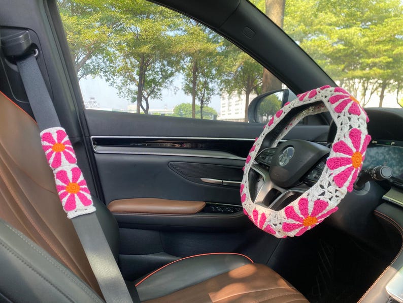 May include: A white crocheted steering wheel cover with pink flowers. The cover is installed on a car's steering wheel. The car's interior is brown leather.