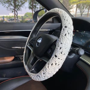 May include: White crocheted steering wheel cover with a floral design. The cover is installed on a black leather steering wheel in a car.