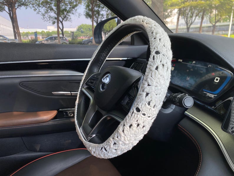 May include: White crocheted steering wheel cover with a floral design. The cover is installed on a black leather steering wheel in a car.