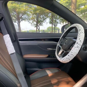 May include: A white crocheted steering wheel cover in a car with brown leather seats. The car is parked and the windows are down, showing a view of trees outside.