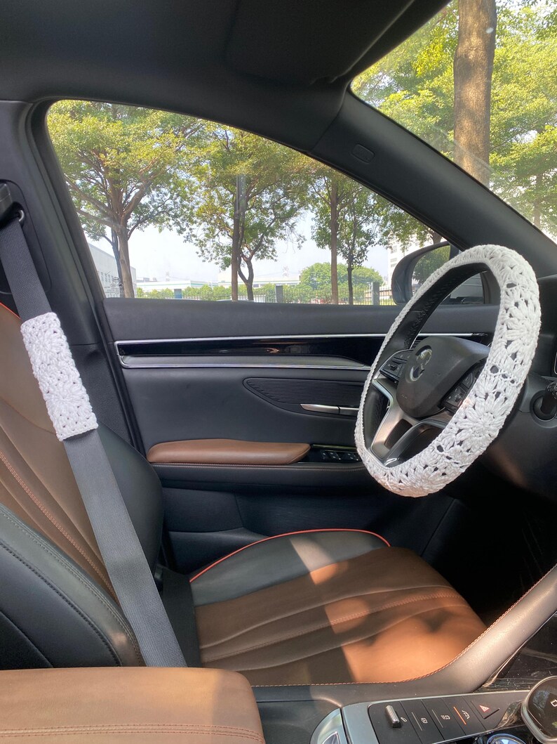 May include: A white crocheted steering wheel cover in a car with brown leather seats. The car is parked and the windows are down, showing a view of trees outside.