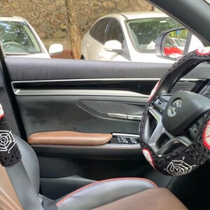 Op de afbeelding: Een zwart auto-interieur met een zwart-witte gehaakte stuurhuls met een spinnenweb- en oogontwerp. De veiligheidsgordel heeft een bijpassende gehaakte hoes met een oogontwerp.
