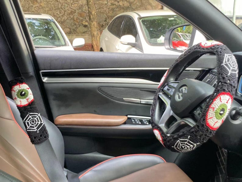 Op de afbeelding: Een zwart auto-interieur met een zwart-witte gehaakte stuurhuls met een spinnenweb- en oogontwerp. De veiligheidsgordel heeft een bijpassende gehaakte hoes met een oogontwerp.