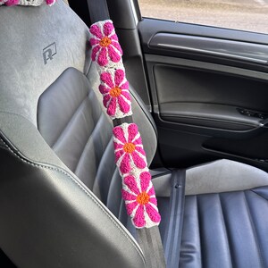 May include: A white car seatbelt with pink and orange crocheted flower embellishments. The seatbelt is in a car with black leather seats.