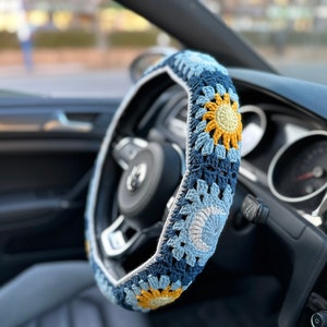 May include: Crocheted steering wheel cover in shades of blue, yellow, and white. The cover features a floral design with a sunflower motif. The cover is on a black steering wheel inside a car.