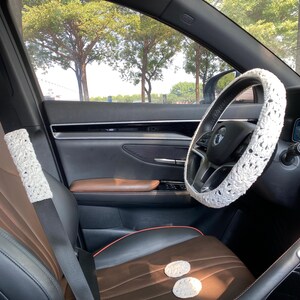 May include: A white crocheted steering wheel cover, a white crocheted seatbelt cover, and two white crocheted coasters in a car interior with brown leather seats.