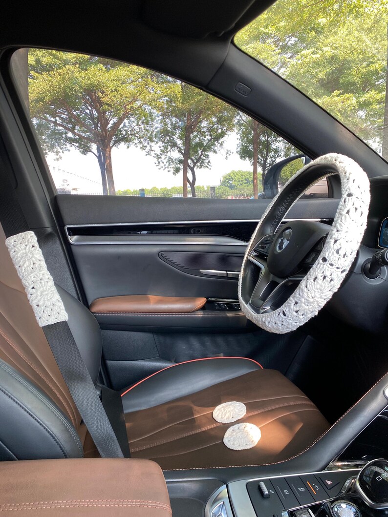 May include: A white crocheted steering wheel cover, a white crocheted seatbelt cover, and two white crocheted coasters in a car interior with brown leather seats.