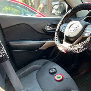 Op de afbeelding: Een zwart auto-interieur met een gehaakte stuurwielhoes en gordelhoes met een spinnenweb- en oogontwerp. De stuurwielhoes is zwart met witte spinnenwebben en een rood oog met een groene iris. De gordelhoes is zwart met een wit spinnenweb en een rood oog met een groene iris. Er zijn twee kleine gehaakte oog- en spinnenweb-patches op de stoel.
