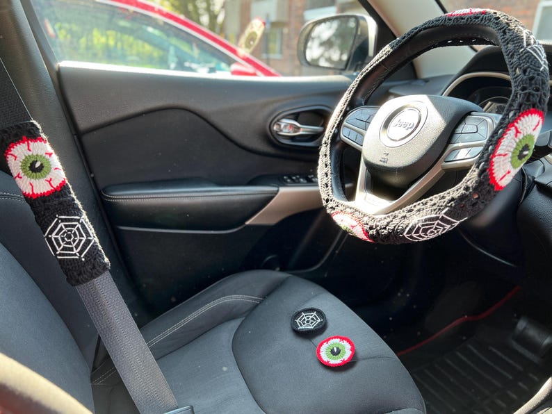 Op de afbeelding: Een zwart auto-interieur met een gehaakte stuurwielhoes en gordelhoes met een spinnenweb- en oogontwerp. De stuurwielhoes is zwart met witte spinnenwebben en een rood oog met een groene iris. De gordelhoes is zwart met een wit spinnenweb en een rood oog met een groene iris. Er zijn twee kleine gehaakte oog- en spinnenweb-patches op de stoel.