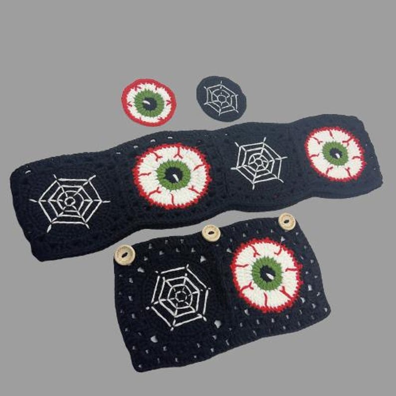 Op de afbeelding: Een set gehaakte Halloween-decoraties met zwarte en witte spinnenwebben en rode, witte en groene ogen. De set bevat een lange banner, twee kleine onderzetters en een kleinere banner met knopen.
