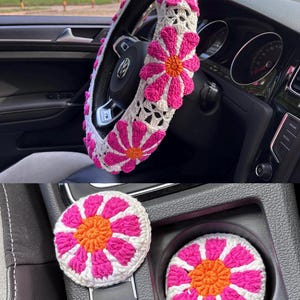 May include: A set of crocheted car accessories in pink and white with orange centers. The set includes a steering wheel cover, a gear shift knob cover, and two cup holders.